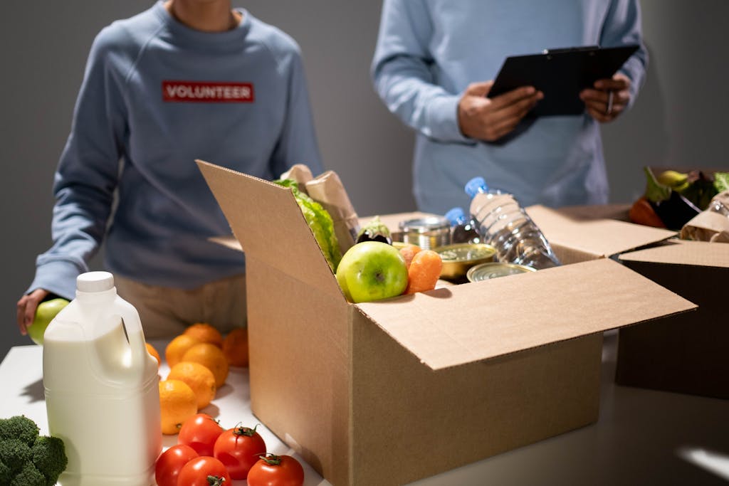 People packing a cardboard box with essentials like fruits, vegetables, and bottled water for charity.